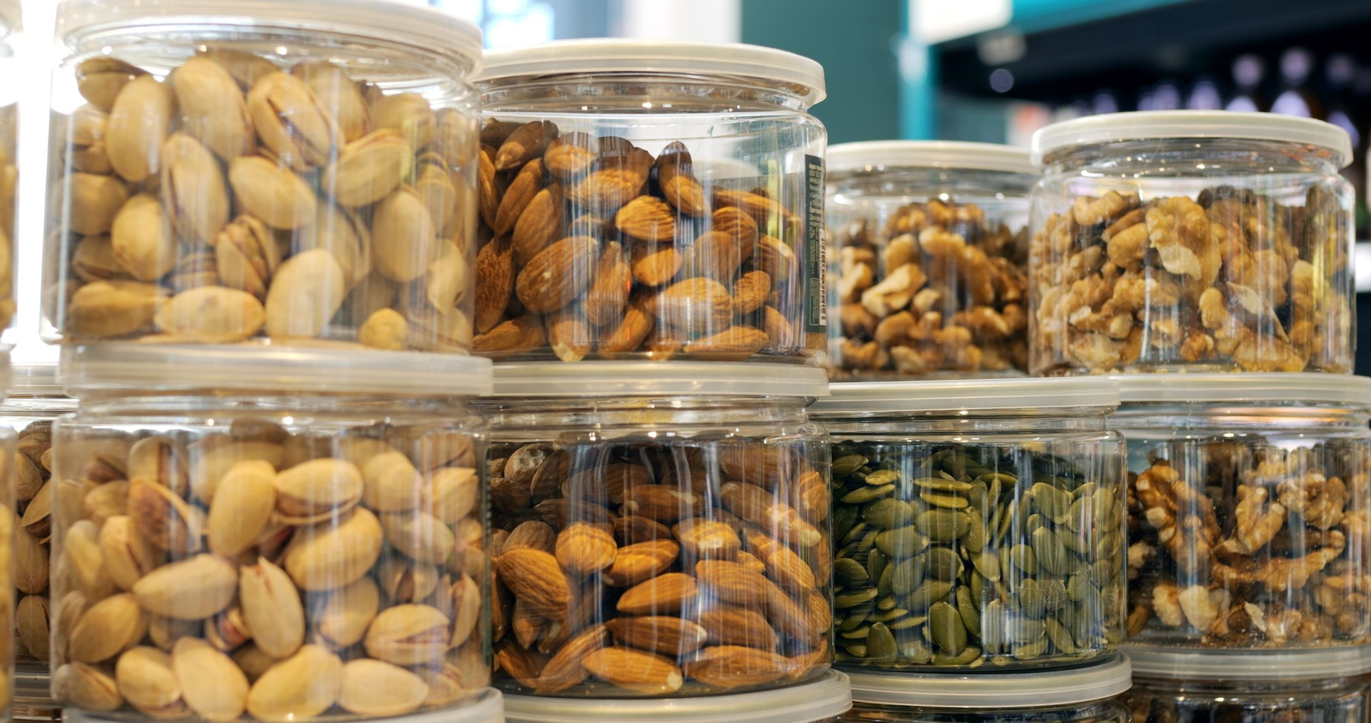 Pistachios, cashews, almonds, walnuts, pumpkin seeds, and other nuts and seeds are neatly arranged in clear plastic containers, creating an appealing display in a grocery store or market setting